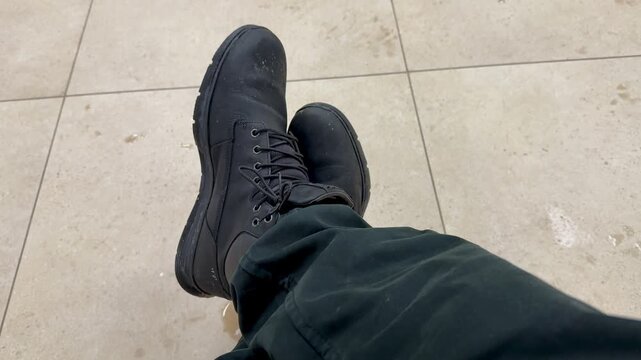 Dirty black boots overhead on tile, worn mens work boots scuffed and dusty, laces tied, dark pants cuff visible, neutral ceramic grout, subtle stain nearby.