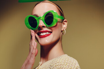 Smiling woman with bright red lipstick wears large green sunglasses and beige sweater. She poses with hand near face on yellow background. Concept of fashion, style, and summer mood.