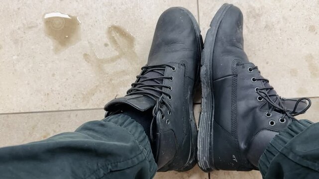 Dirty black boots overhead on tile, worn mens work boots scuffed and dusty, laces tied, dark pants cuff visible, neutral ceramic grout, subtle stain nearby.