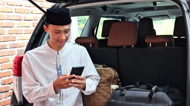 An Asian Muslim man sits in the trunk of a car typing a message on his cell phone.