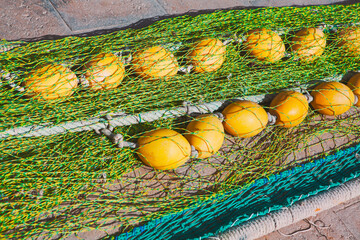 Fishing net with yellow spherical floats is laid out on pavement, held together by green mesh and thick ropes. Aquatic netting equipment, specifically the floats and twine