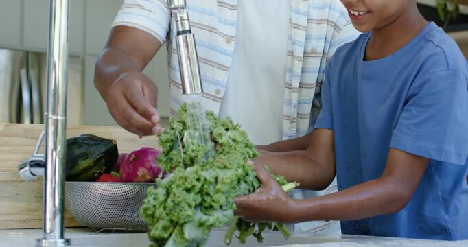 African American father and son turning faucet holding kale and rinsing to clean at kitchen sink