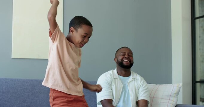 African American father and teen son gaming on couch, son standing, celebrating controller win