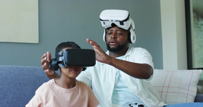 African American dad and son on couch son placing VR headset dad adjusting guiding with controller