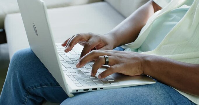 Person is placing white laptop on lap and is typing on keyboard while working on couch