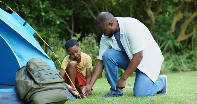 African American dad and boy pitching tent, securing guyline and stake with mallet in yard