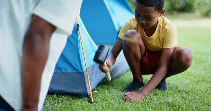 African American father and son securing pitched tent, child tapping stake with mallet to anchor