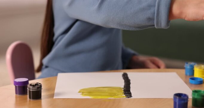 Little girl drawing with brush and paints at wooden table indoors, closeup. Camera moving left