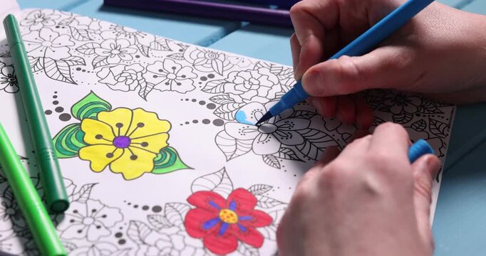 Woman coloring antistress picture with felt tip pen at light blue wooden table, closeup
