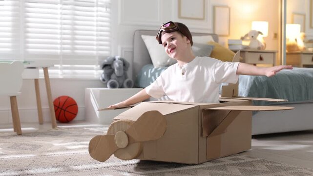 Happy little boy in cardboard airplane and goggles playing pilot on floor at home. Camera moving forward and backwards