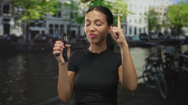 Hispanic brunette woman holding a handgrip exerciser and finger pointing up on a street by an amsterdam canal, bicycles beside her; confidence empowerment concentration.