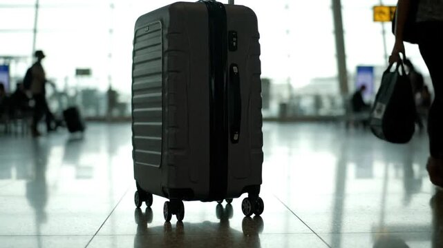 A close-up shot of a hard-shell suitcase standing on the shiny floor of an airport terminal, with travelers passing by in the background and natural daylight illuminating the scene.