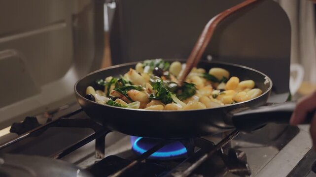 Woman cooking gnocchi with vegetables in a frying pan on a gas stove in a camper van in slow motion