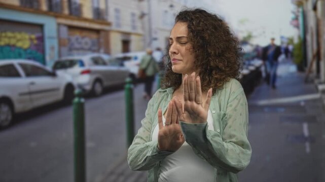 Woman pressing both palms forward in a stop gesture on street near parked cars and bollards, visible hands and shoulders in urban sidewalk scene; unease boundary.