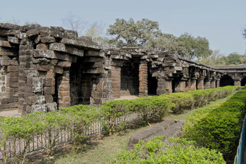 ​a perspective view of the outer circular corridor at kurumbera fort, showcasing heavy laterite stone pillars, weathered masonry, and a well-maintained garden path under a clear afternoon sky