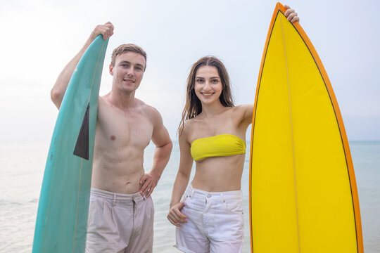 couple standing with surfboards in shallow ocean under cloudy sky. Summer surfing lifestyle, tropical beach vacation, travel adventure and active outdoor recreation concept.