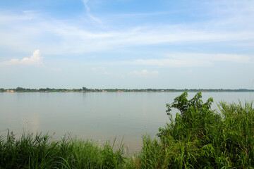 Peaceful wide river with lush green banks and distant village under a calm blue sky, evoking serene natural landscape.