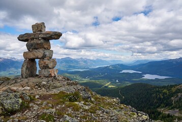 Stone inukshuk marker on rocky mountain summit with scenic view of forest valley and distant lakes