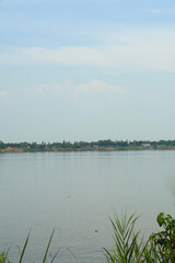Serene wide river with calm water reflecting soft blue sky, showcasing distant lush green tropical trees along the quiet bank.
