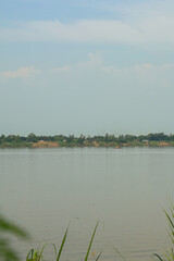Peaceful wide river reflecting soft blue sky, distant green bank with tropical trees and subtle buildings, evoking serene natural beauty.