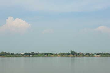 Panoramic view of a calm wide river with distant lush green tropical riverbank under a soft blue sky and white clouds.