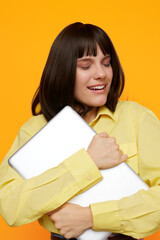 Fototapeta premium Bright portrait of a cheerful woman hugging a laptop, wearing a soft yellow shirt, with a warm orange background and a joyful, relaxed mood.