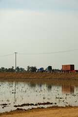 Heavy semi-truck transports orange shipping container on rural road next to flooded rice paddy field under bright sky.