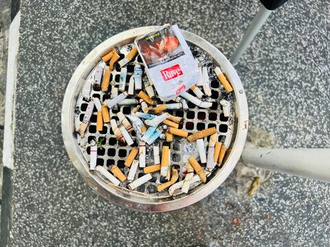 Sarawak, Malaysia - 15 February 2026: High-angle close-up of a circular outdoor metal cigarette ashtray filled with numerous extinguished cigarette butts and a discarded white "Rave" brand cigarette.