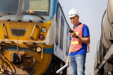 Railway engineer using walkie-talkie for coordinating freight transport and logistics at train station, Worker in safety vest and hardhat conducting inspection with radio communication near locomotive