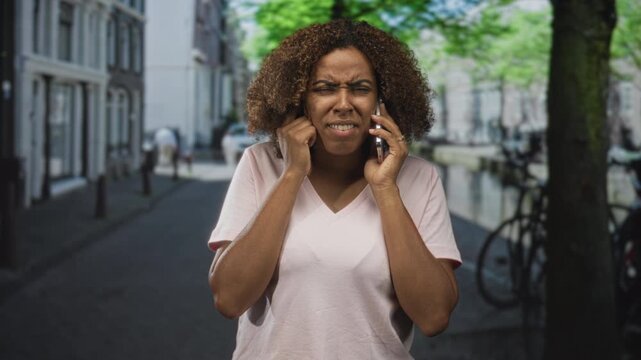 Woman holding smartphone to ear and plugging other ear with finger on city street near canal; frustration delay.