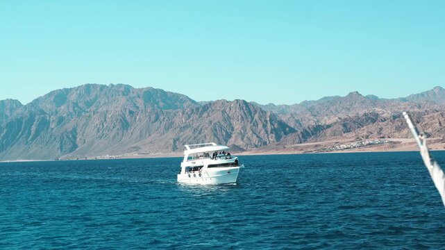 A luxury white yacht and passenger ferry cruise through the blue water of the summer sea toward a tropical island harbor within the rocky mountains on a red sea of the gulf of agaba dahab egypt