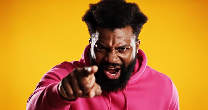 Angry man in a pink hoodie pointing at the camera against a bright yellow background