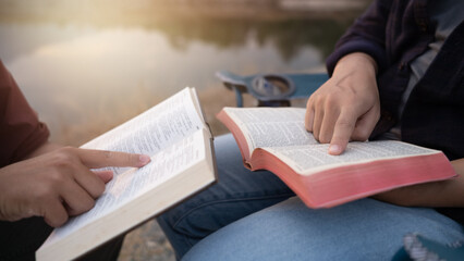 Two people sitting and studying the Holy Bible together outdoors with natural morning sunlight, close up of hand pointing at scripture.
