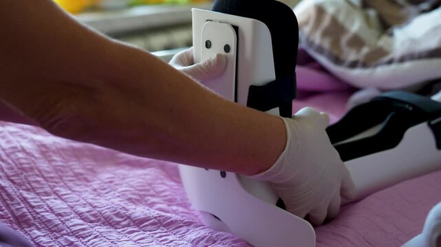 Healthcare Professional Applying Orthopedic Foot Brace to Patient in Bed. Caregiver hands adjusting a medical foot immobilizer or splint on a patient. Concept of rehabilitation, orthopedic care