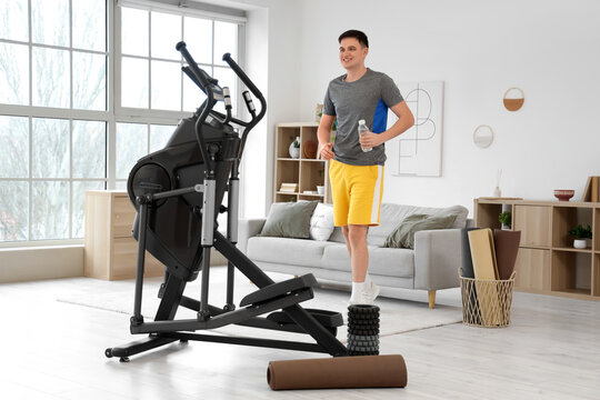 Sporty young man with water bottle walking to modern exercising bike at home