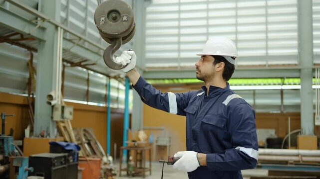 Professional male engineer in a hard hat operating an industrial overhead crane with a remote control inside a large warehouse factory. Focused worker managing heavy lifting and safety equipment.