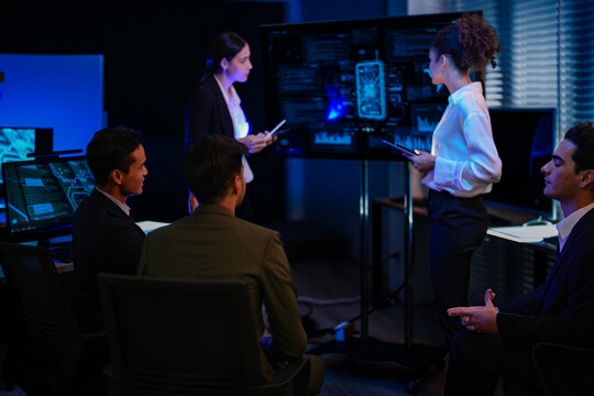Professional IT team analyzing global network security dashboard in modern control center. Female manager leading cyber technology project discussion in dark night office.