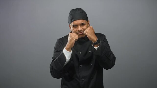 Hispanic chef man with beard wearing black chef jacket and headscarf raises fists and punches in a studio, hands close to face; determination focus strength.