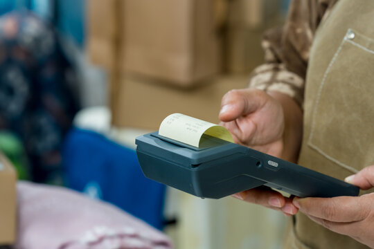 Senior asian retired businesswoman holds POS terminal, prints receipt, checks paperwork order processing, verifies transaction total, prepares parcel packaging for small business online shipping.