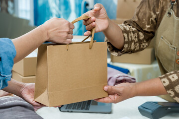 Senior Asian businesswoman entrepreneur gives kraft paper bag pickup to adult Asian female customer, verifies order invoice receipt, checks barcode label, carton stack near laptop in studio workshop. © Ekkasit A Siam