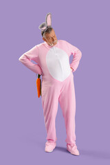 Senior man in Easter bunny costume with plush carrot on lilac background