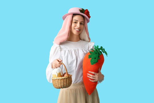 Beautiful young woman in bunny ears holding toy carrot and basket with Easter bread on blue background. Easter celebration