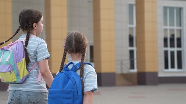 concept schoolchildren, child go holding hands school backpacks, friends team, child walk through school yard backpacks, happy team classmates, view from back, girls pigtails go to class together