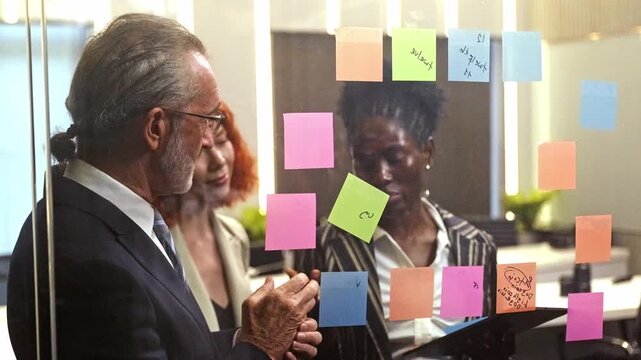 Diverse team collaborating in a glass-walled office, discussing ideas around colorful sticky notes during a planning meeting.