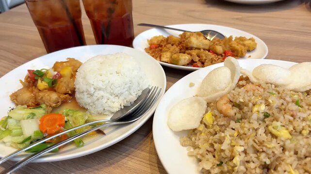 White rice, sweet‑sour chicken, shrimp fried rice, stir‑fried vegetables, and spicy fried bites are arranged neatly on a table beside two iced tea glasses
