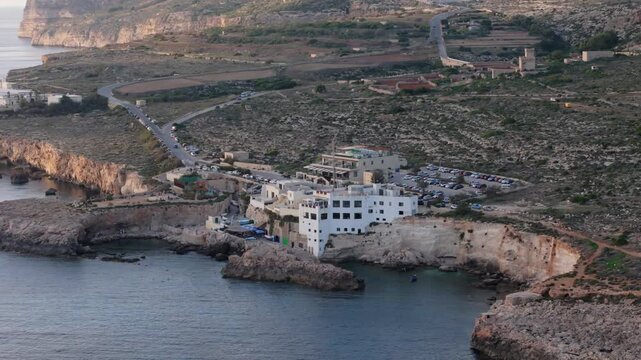 Slow aerial pullback revealing Ghar Lapsi on the southern coast of Malta before storm damage. Calm waters intact limestone cliffs nearby roads and coastal buildings are shown in warm evening light.