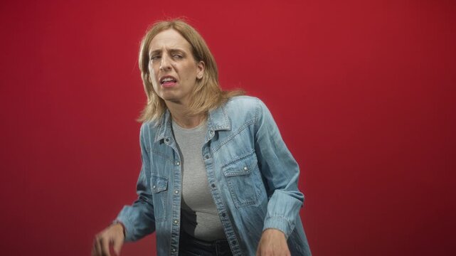Woman cupping hand to ear while leaning forward with a puzzled frown, wearing a denim jacket and grey tshirt against a studio red backdrop; confusion curiosity.