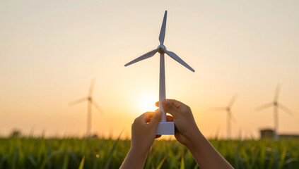 Hands holding a miniature model of a wind turbine against a backdrop of wind turbines in a grassy field at sunset.
