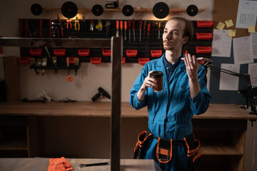 Fototapeta premium Handyman in blue overalls resting drinking coffee and communicating with AI assistant in workshop
