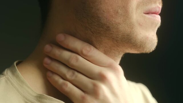 Unshaven man experiencing throat pain, touching his neck in discomfort against dark background. Close up shot showing symptoms of flu, tonsillitis, or other respiratory infections